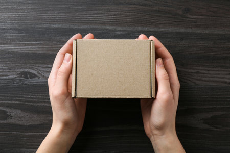 Woman with blank cardboard box on dark wooden background, top view. Mockup for design