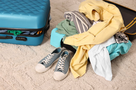 Suitcase with pile of different messy clothes, another one and sneakers on carpetの写真素材