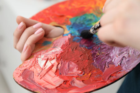 Woman with paintbrush mixing paints on palette against blurred background, closeupの写真素材