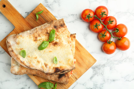 Halves of tasty calzone, basil, and tomato on white marble table, flat lay
