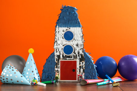 Bright pinata in shape of spaceship and party accessories on table against orange background