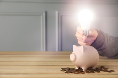 Energy saving concept. Man holding glowing light bulb over piggy bank at wooden table, closeup. Space for textの写真素材