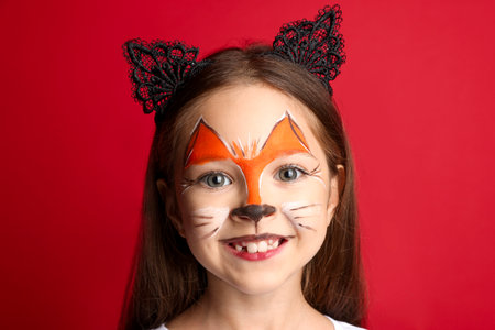 Cute girl with painted face and ears as cat on red background, closeup