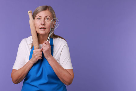 Surprised woman with rolling pin and whisk on violet background. Space for textの写真素材