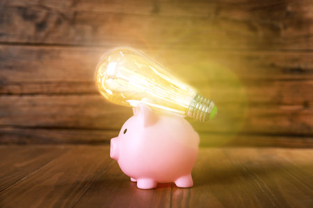 Piggy bank and glowing light bulb on wooden table, space for text. Energy saving conceptの写真素材