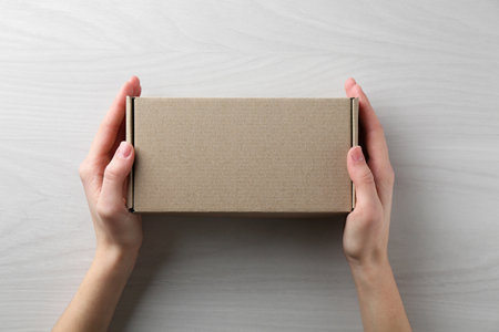 Woman with blank cardboard box on white wooden background, top view. Mockup for designの写真素材