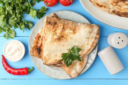 Halves of tasty calzone, tomato, chili pepper, parsley and sauce on light blue table, flat lay