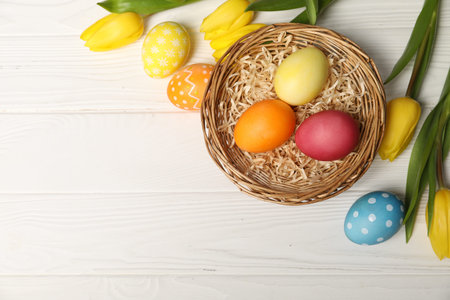Easter eggs in wicker basket and beautiful tulips on white wooden table, flat lay. Space for text