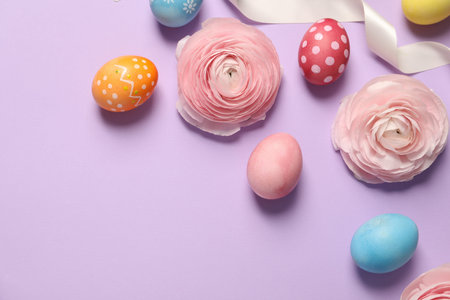 Easter eggs, ranunculus flowers and ribbon on lilac background, flat lay. Space for text