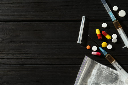 Drug addiction. Plastic bag with powder, syringes and pills on black wooden table, flat lay. Space...