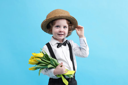 Cute little boy in wicker hat with bouquet of tulips on light blue background