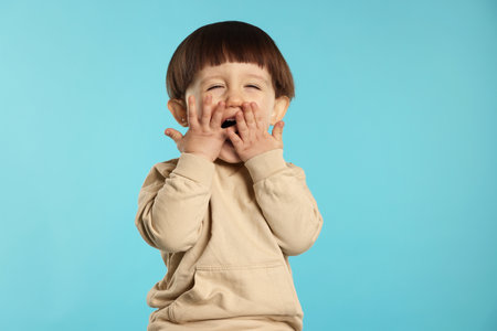 Portrait of emotional little boy covering mouth on light blue backgroundの写真素材