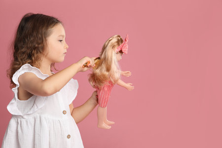 Cute little girl brushing doll's hair on pink background. Space for textの写真素材