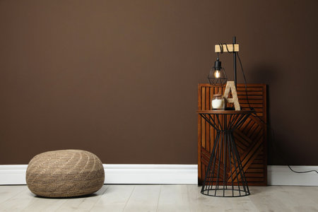 Comfortable knitted pouf and table with decor elements near brown wall indoors, space for text. Interior designの写真素材