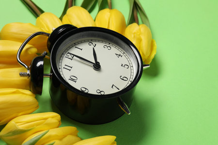 Spring time. Alarm clock and beautiful tulips on green background, closeup. Space for text