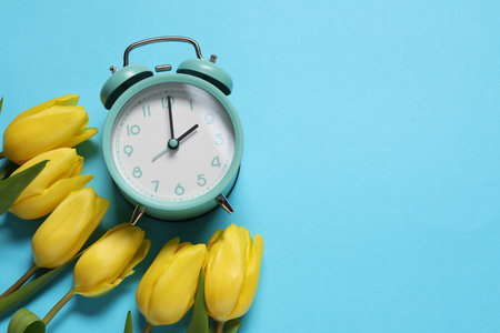 Spring time. Alarm clock and beautiful tulips on light blue background, flat lay. Space for textの写真素材