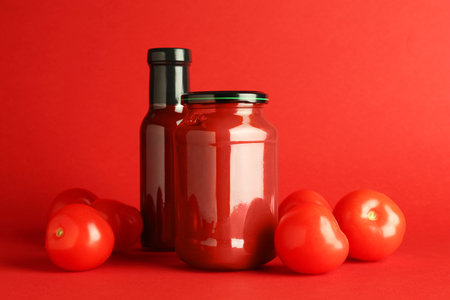 Ketchup in glass bottle, jar and fresh tomatoes on red backgroundの写真素材