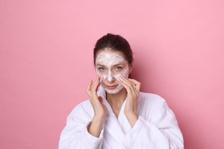 Spa day. Beautiful woman applying mask onto her face on pink background