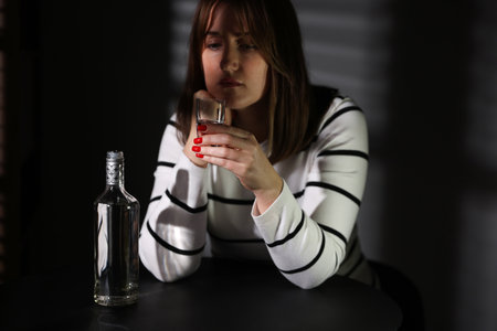 Alcohol addiction. Woman with shot glass of vodka and bottle at table indoorsの写真素材