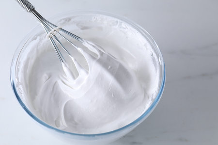 Bowl with whisk and whipped cream on white marble table, closeup