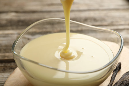 Pouring tasty condensed milk into bowl on wooden table, closeupの写真素材