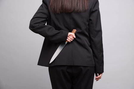 Businesswoman with knife behind her back on light grey background, closeup
