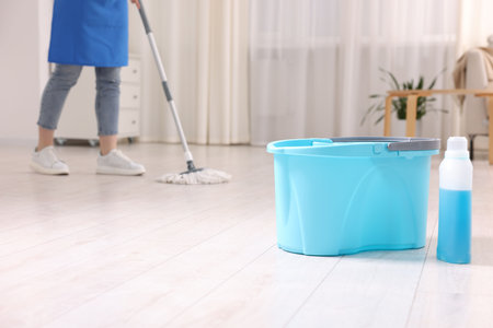 Woman floor cleaning with string mop indoors, focus on bucket and bottle of detergentの写真素材