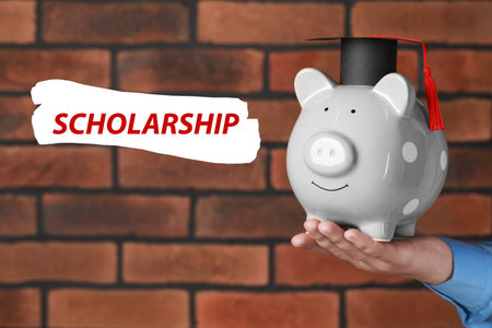 Scholarship. Man holding piggy bank with graduate hat against brick background, closeupの写真素材