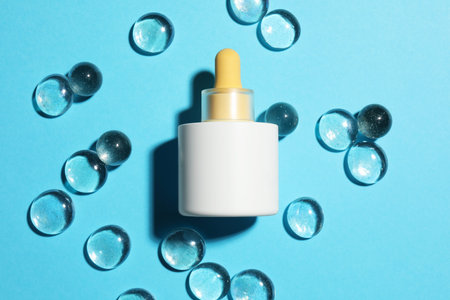 Bottle of skin care product and transparent decorative stones on light blue background, flat lay