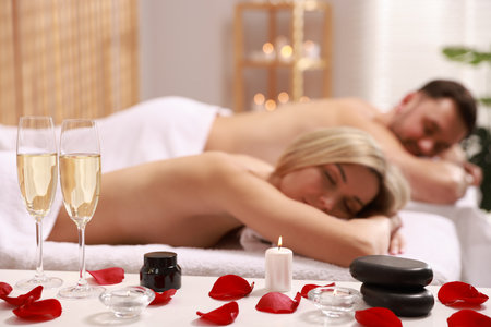 Couple lying on massage tables in spa salon, focus of petals, burning candle and glasses of champagneの写真素材