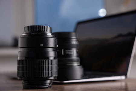 Professional photo camera lenses and laptop on wooden desk, closeup