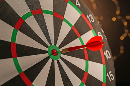 Red arrow hitting target on dart board against black background with blurred lights, closeupの写真素材
