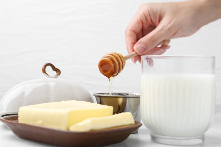 Woman with honey dipper, closeup. Fresh milk in glass and butter on white tiled tableの写真素材