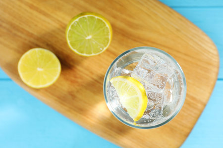 Refreshing soda water with ice cubes in glass, cut lemon and board on light blue wooden table, top...