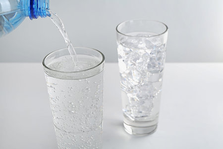 Pouring refreshing soda water into glass at white table against light background