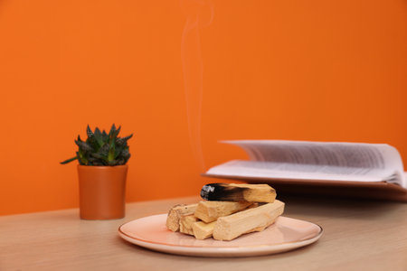 Palo santo sticks, book and houseplant on wooden table against orange wall, space for textの写真素材