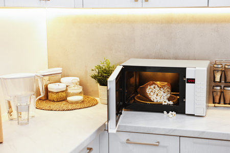 Open microwave oven with bag of popcorn on white marble countertop in kitchenの写真素材