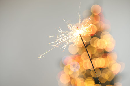 Bright burning sparkler on gray background with blurred lights, closeup. Bokeh effect