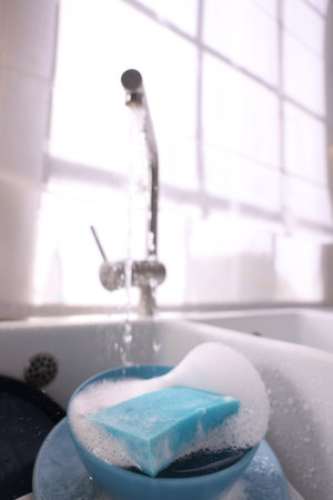Dishes, blue sponge and foam under tap water in kitchen sink, closeupの写真素材