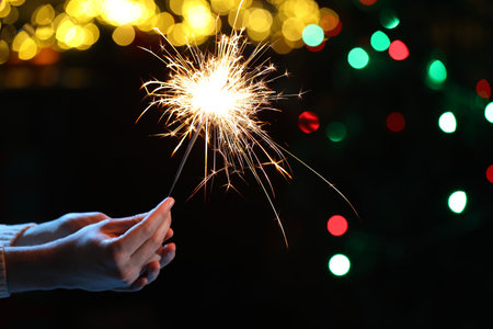 Man holding bright burning sparkler on dark background with blurred lights, closeup. Bokeh effectの写真素材
