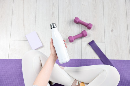 Woman with thermo bottle, dumbbells and planner on floor indoors, top view
