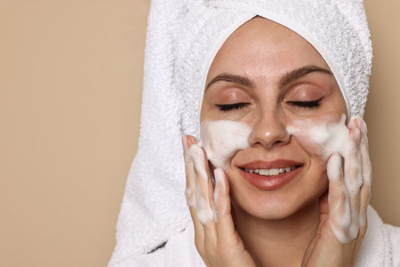 Beautiful woman washing her face with cleansing foam on beige background, space for textの写真素材