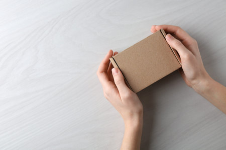 Woman with blank cardboard box on white wooden background, top view. Mockup for design
