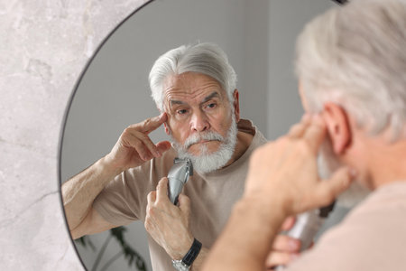 Senior man trimming beard near mirror in bathroomの写真素材