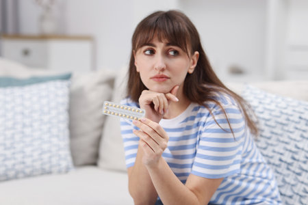 Upset woman with blister of contraceptive pills on sofa at homeの写真素材