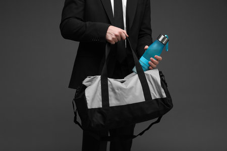 Man in suit putting bottle of water into gym bag on dark grey background, closeupの写真素材