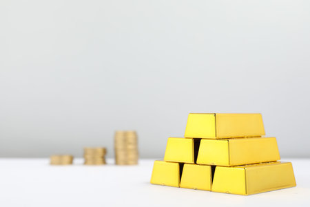 Shiny gold bars and coins on white table, space for text