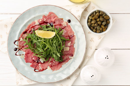 Delicious beef carpaccio with arugula, cheese, lemon and capers on white wooden table, flat layの写真素材