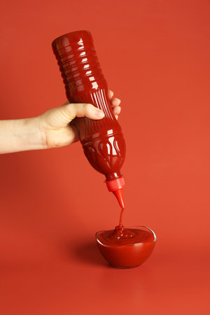 Woman squeezing ketchup from bottle into bowl on red background, closeupの写真素材