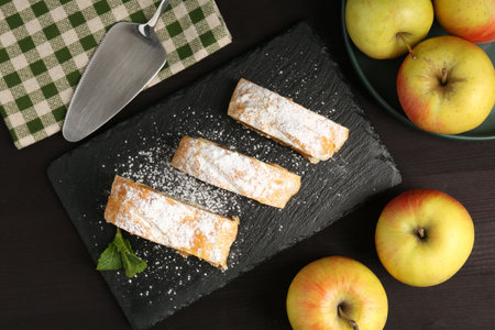 Pieces of tasty apple strudel with powdered sugar, mint and fruits on black wooden table, flat layの写真素材